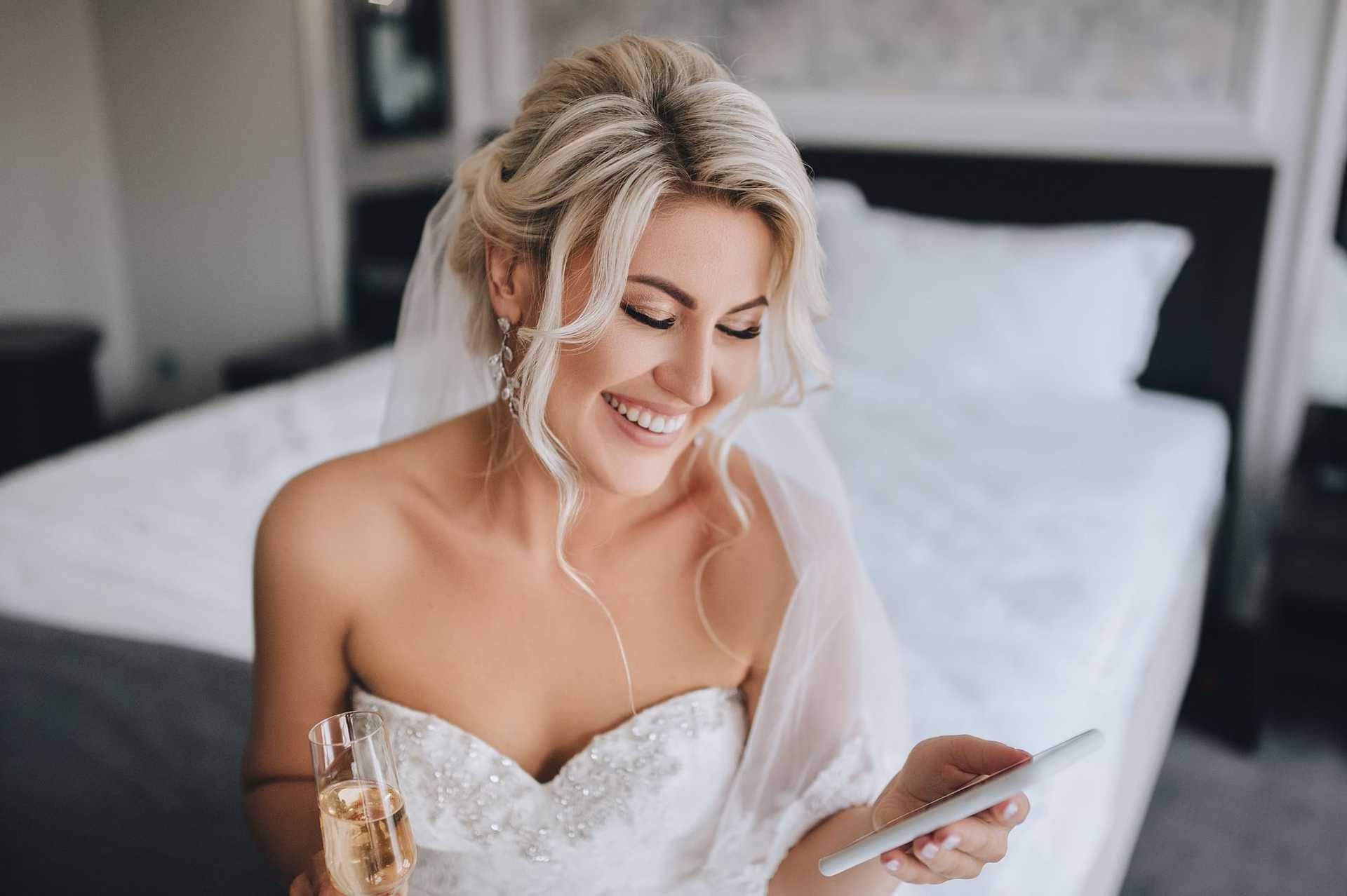 Bride in a wedding dress smiles while holding a phone and a glass of champagne.